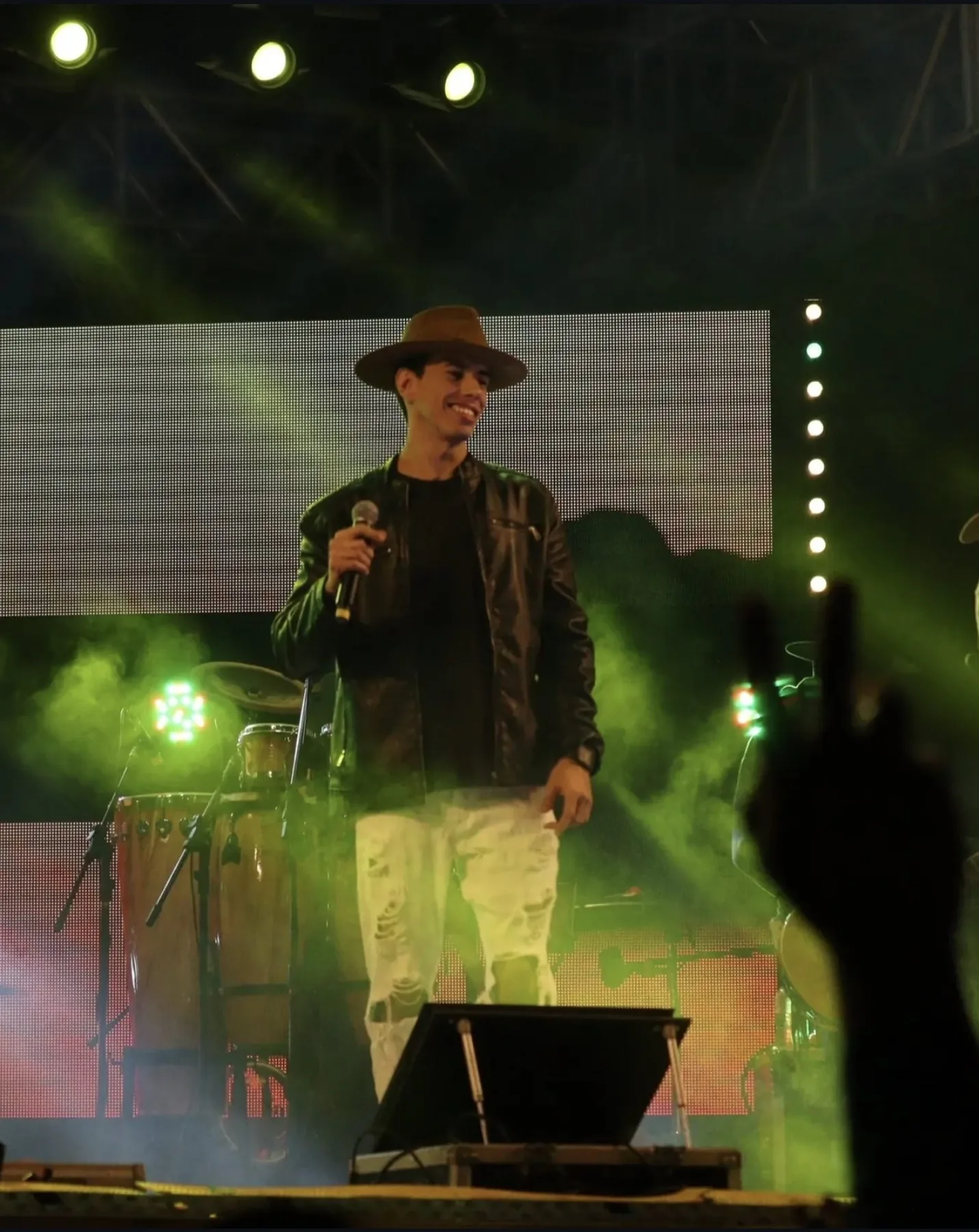 Anderson Junior sorrindo no palco entre luzes verdes, durante apresentação
